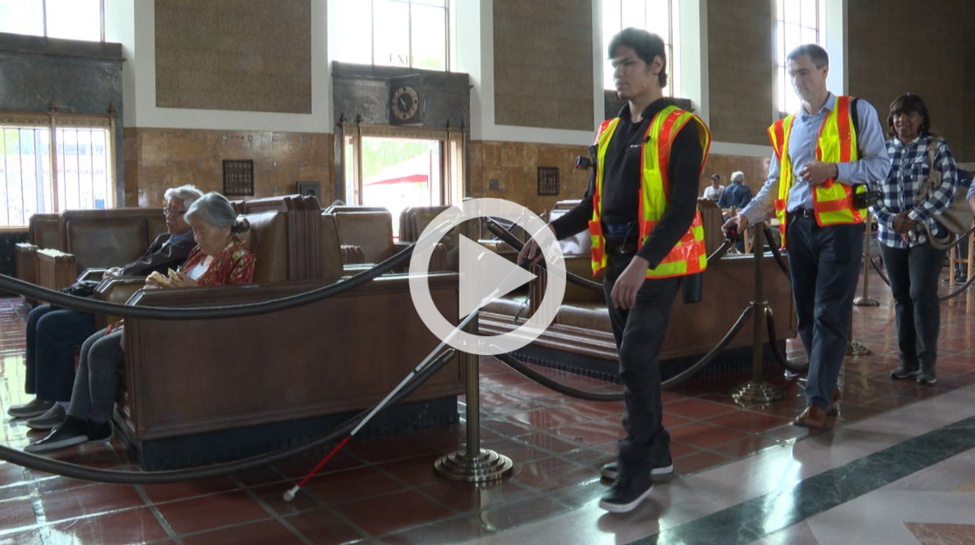 Foreground video play button graphic, background person using white cane to navigate through LA Union Station.